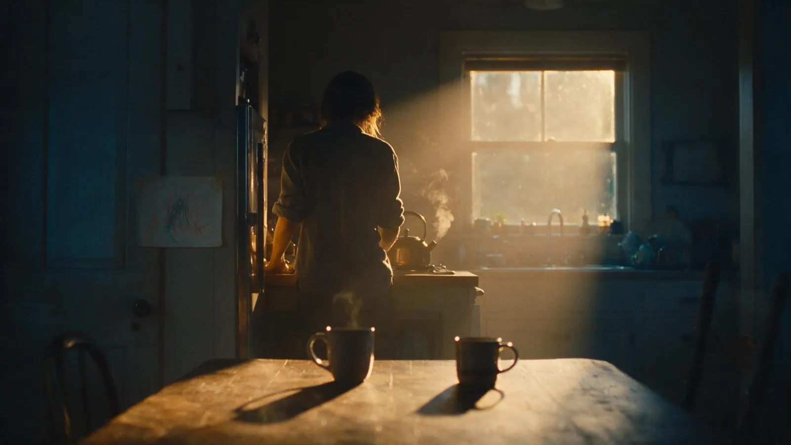 A woman at a kitchen counter in late golden light, steam rising from a kettle, two mugs on the table.